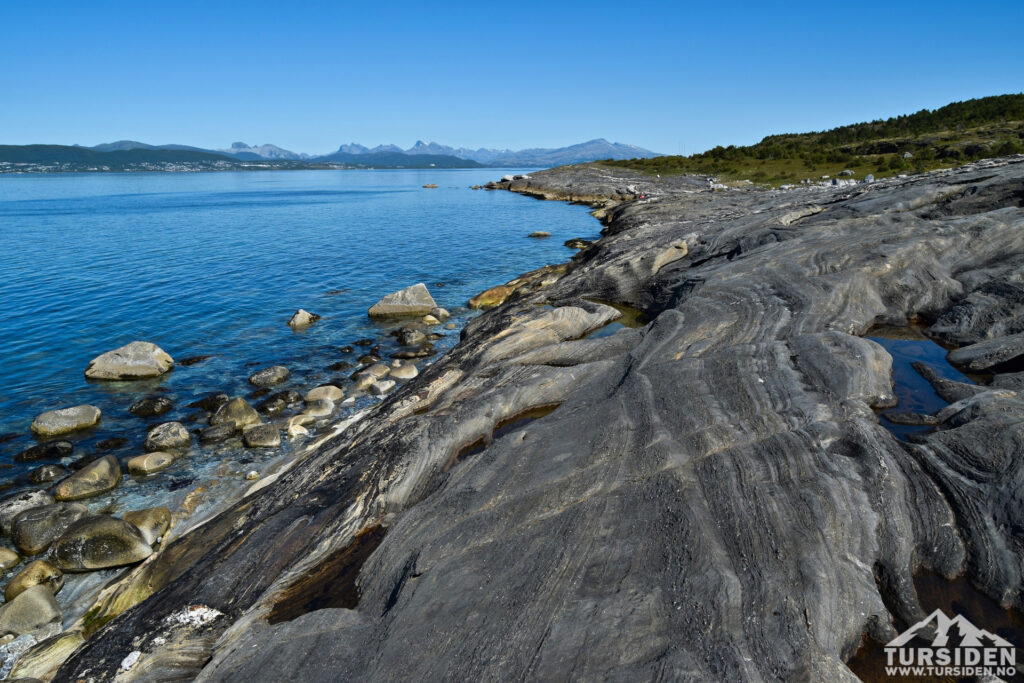 Gestreifte dunkle Felsplatten fallen ins ruhige, blaue Fjord; runde Steine im Wasser, ferne Berge im Sonnenschein.