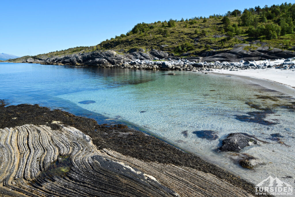 Klare türkisfarbene Bucht mit weißem Sand, gestreiften Felsrippen im Vordergrund und grünem Hügel im Hintergrund.