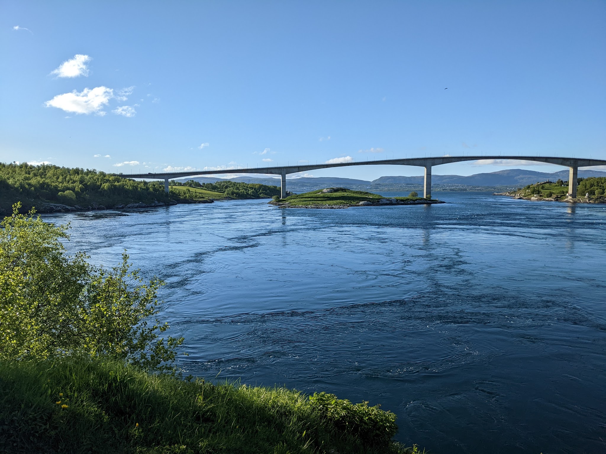 Lange Brücke über blaues, strudelndes Wasser, umgeben von grünen Ufern und fernen Bergen unter klarem Himmel