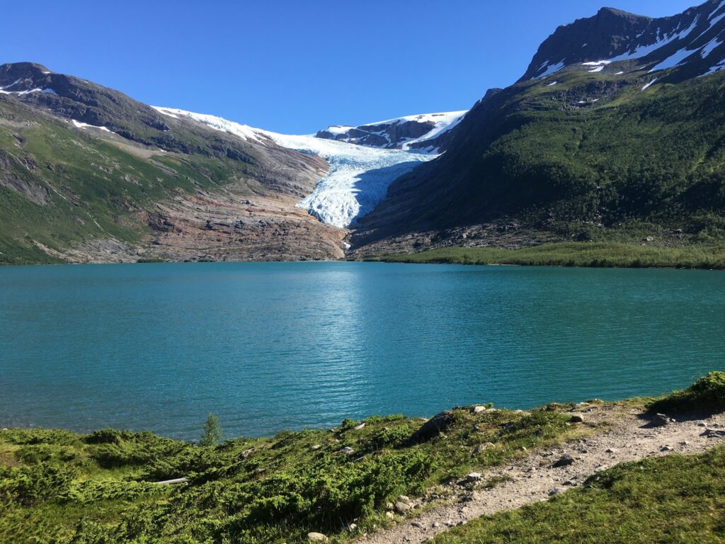 Türkisblauer See vor grünen Bergen, zentraler Gletscher fließt ins Wasser, klarer Himmel.
