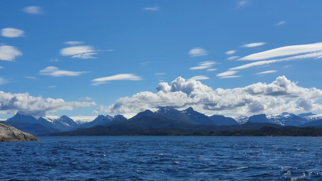 Tiefblaues Meer vor schneebedeckter Bergkette unter sonnigem Himmel mit weißen Schäfchenwolken.
