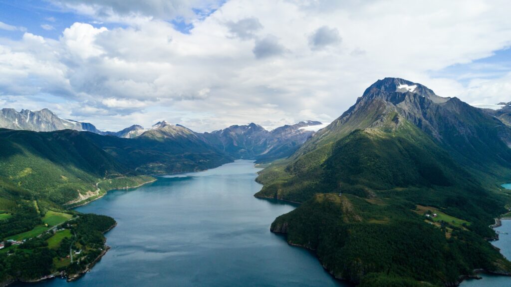 Luftaufnahme eines tiefblauen Fjords, eingerahmt von steilen, bewaldeten Bergen mit felsigen, teils verschneiten Gipfeln