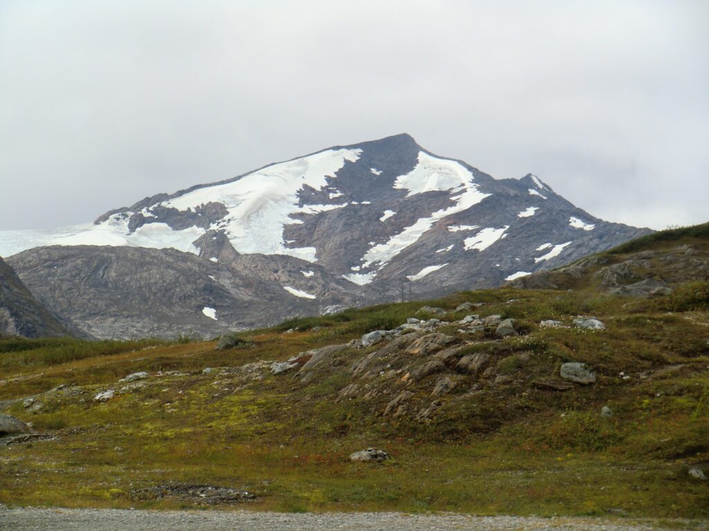 Gletscherbedeckter, felsiger Berggipfel unter grauem Himmel; vorn tundraartige Wiese mit Geröll.