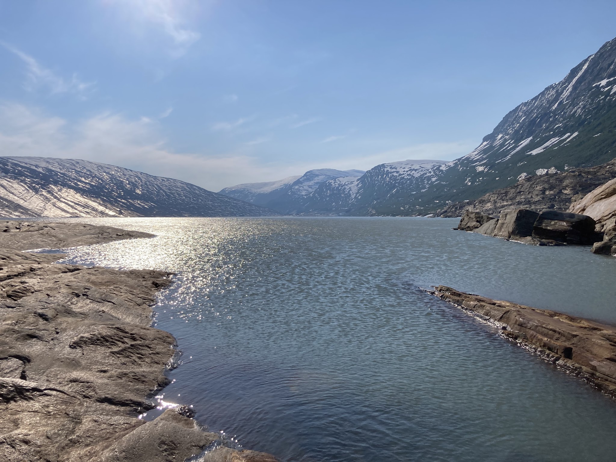 Sonnenglitzerndes Gletscherseeufer zwischen schneebedeckten Bergen unter klarem blauem Himmel.