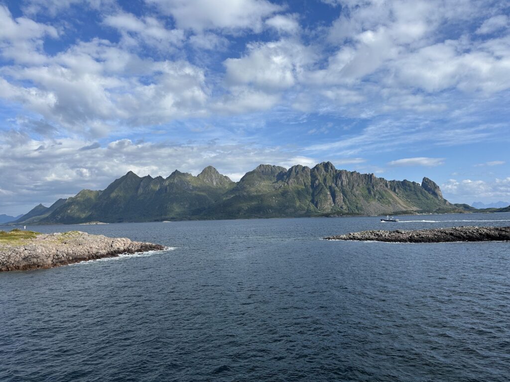 Zackige, grün bewachsene Berge hinter ruhigem Fjord; kleines Boot fährt an zwei steinigen Landzungen vorbei.