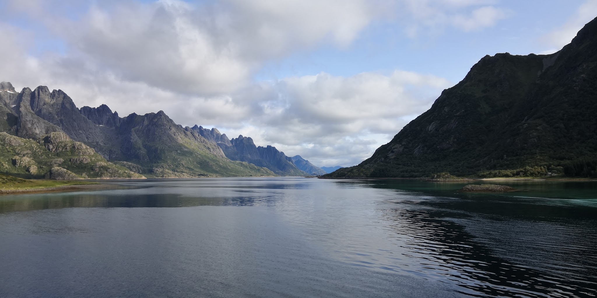 Ruhiger Fjord mit spiegelndem Wasser, flankiert von steilen, grünen Bergen unter teils bewölktem Himmel.