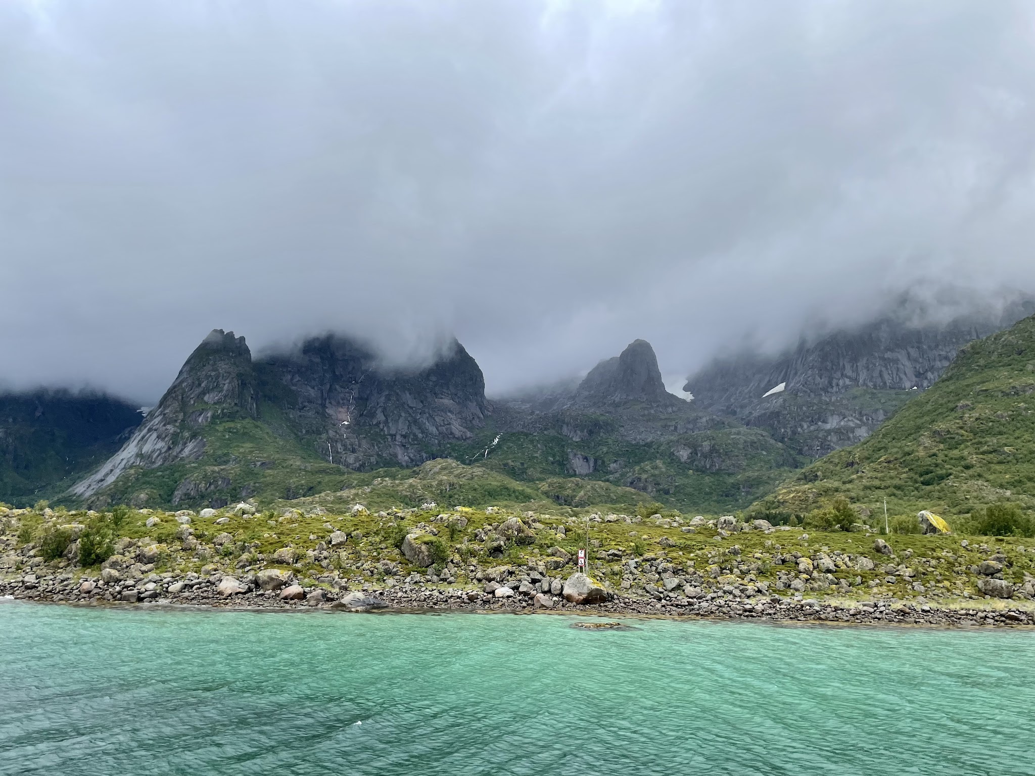 Türkisfarbener Fjord vor grün bewachsenen Felsen und wolkenverhangenen, schroffen Bergspitzen.