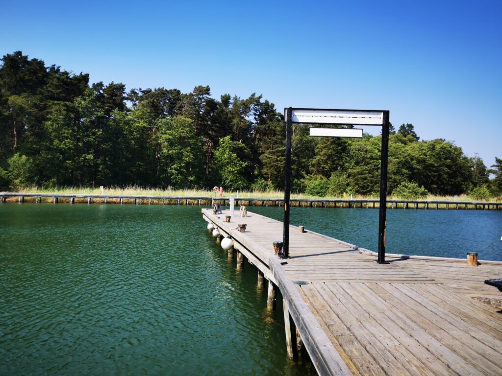 Langer hölzerner Steg führt über grünes Wasser zu bewaldetem Ufer unter wolkenlosem, blauem Himmel.