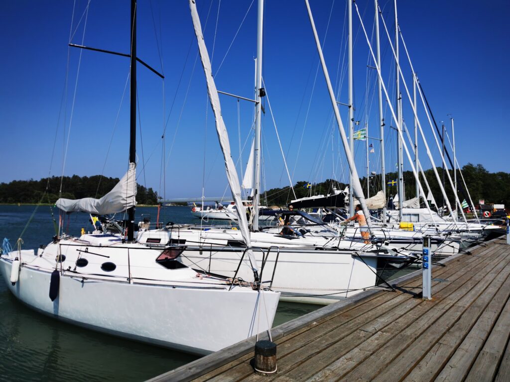 Mehrere weiße Segelboote liegen an einem Holzsteg im sonnigen Hafen; blauer Himmel und schwedische Flaggen im Hintergrund.