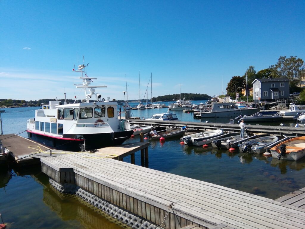 Sonniger Hafen mit weißem Passagierboot am Steg, umgeben von mehreren kleinen Motorbooten auf ruhigem Wasser.