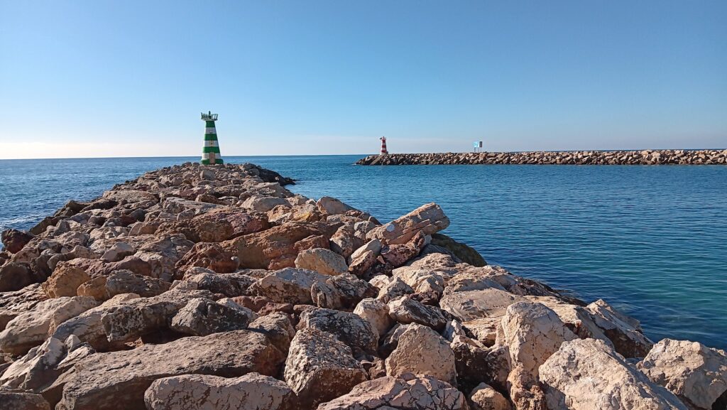 Felsdamm führt zum grün-weiß gestreiften Leuchtturm; ruhiges, blaues Meer, ferne rote Bake, wolkenloser Himmel