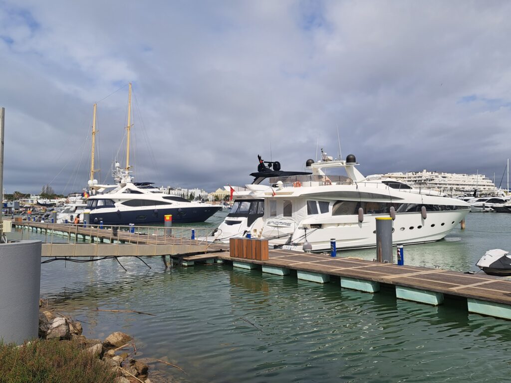 Zwei große Motoryachten liegen am Steg eines Yachthafens unter bewölktem Himmel; grünliches Wasser im Vordergrund.