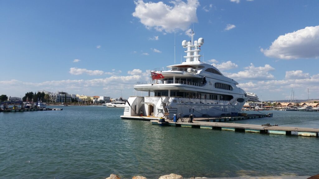 Weiße Luxusyacht liegt an einem Steg in sonnigem Hafen, ruhiges Wasser, niedrige Küstengebäude hinten.