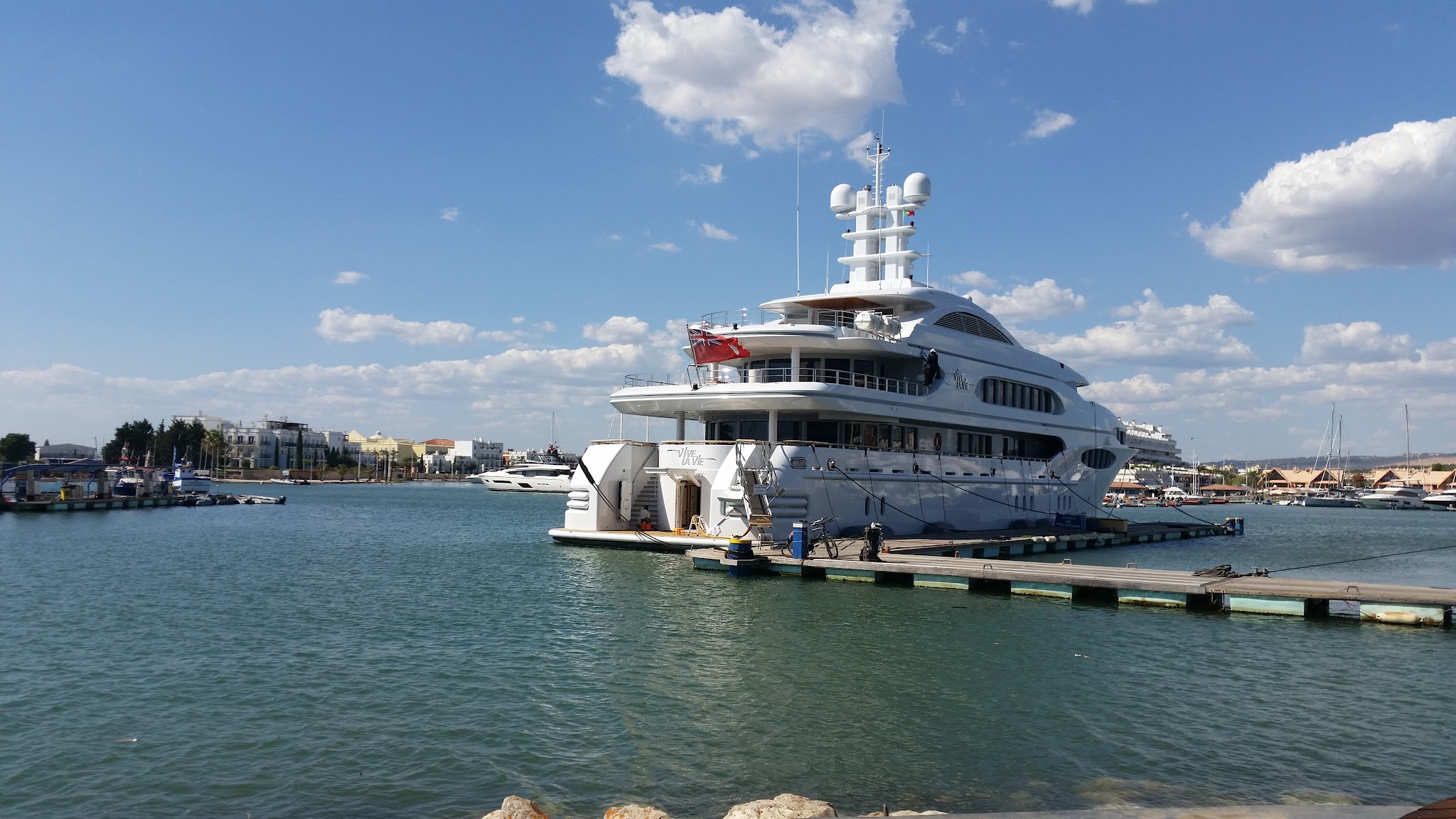 Weiße Luxusyacht liegt an einem Steg in sonnigem Hafen, ruhiges Wasser, niedrige Küstengebäude hinten.