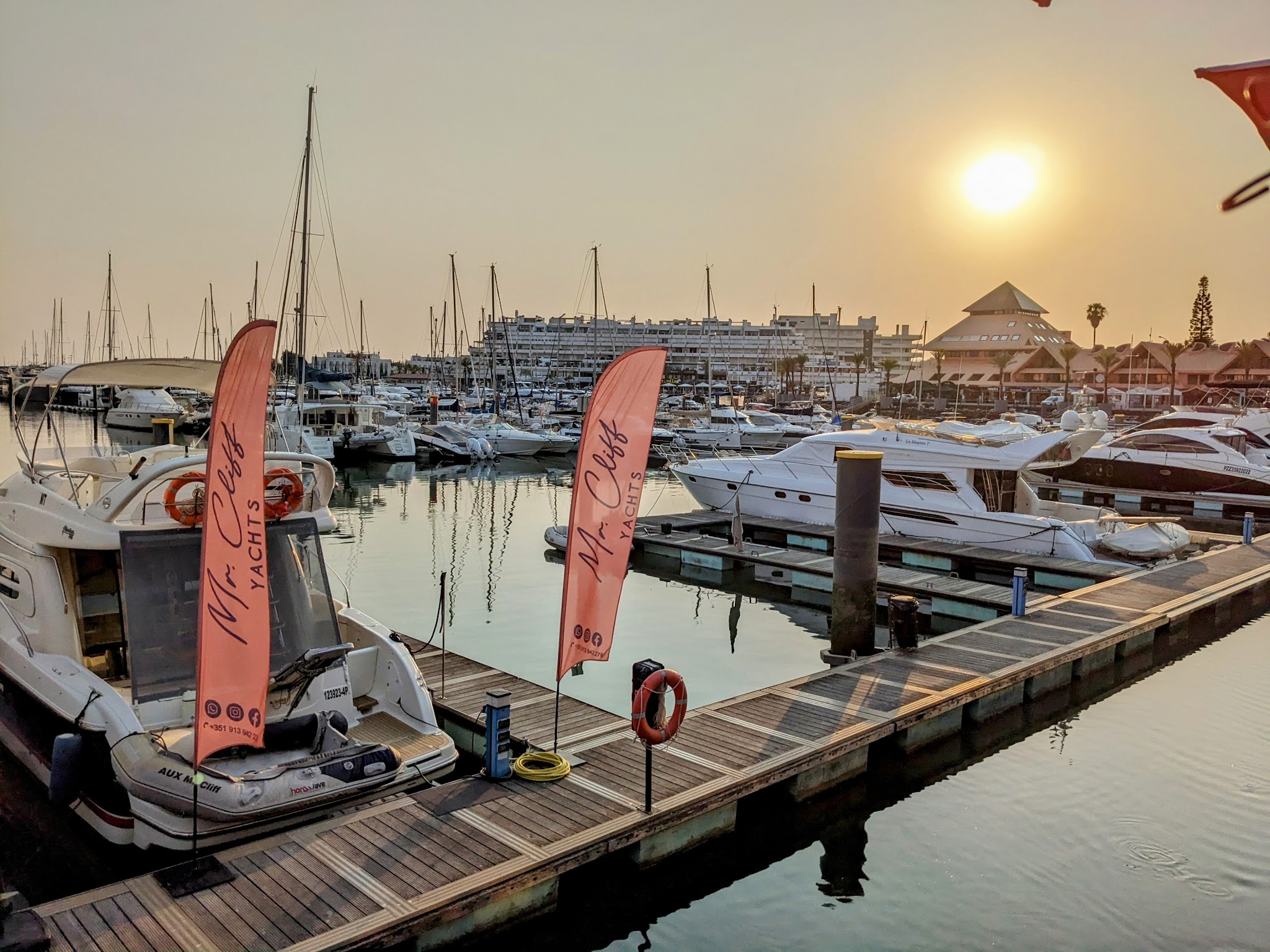 Sonnenuntergang über Yachthafen; Steg mit zwei „Mr. Cliff Yachts“-Fahnen, ringsum angelegte Motor- und Segelboote.