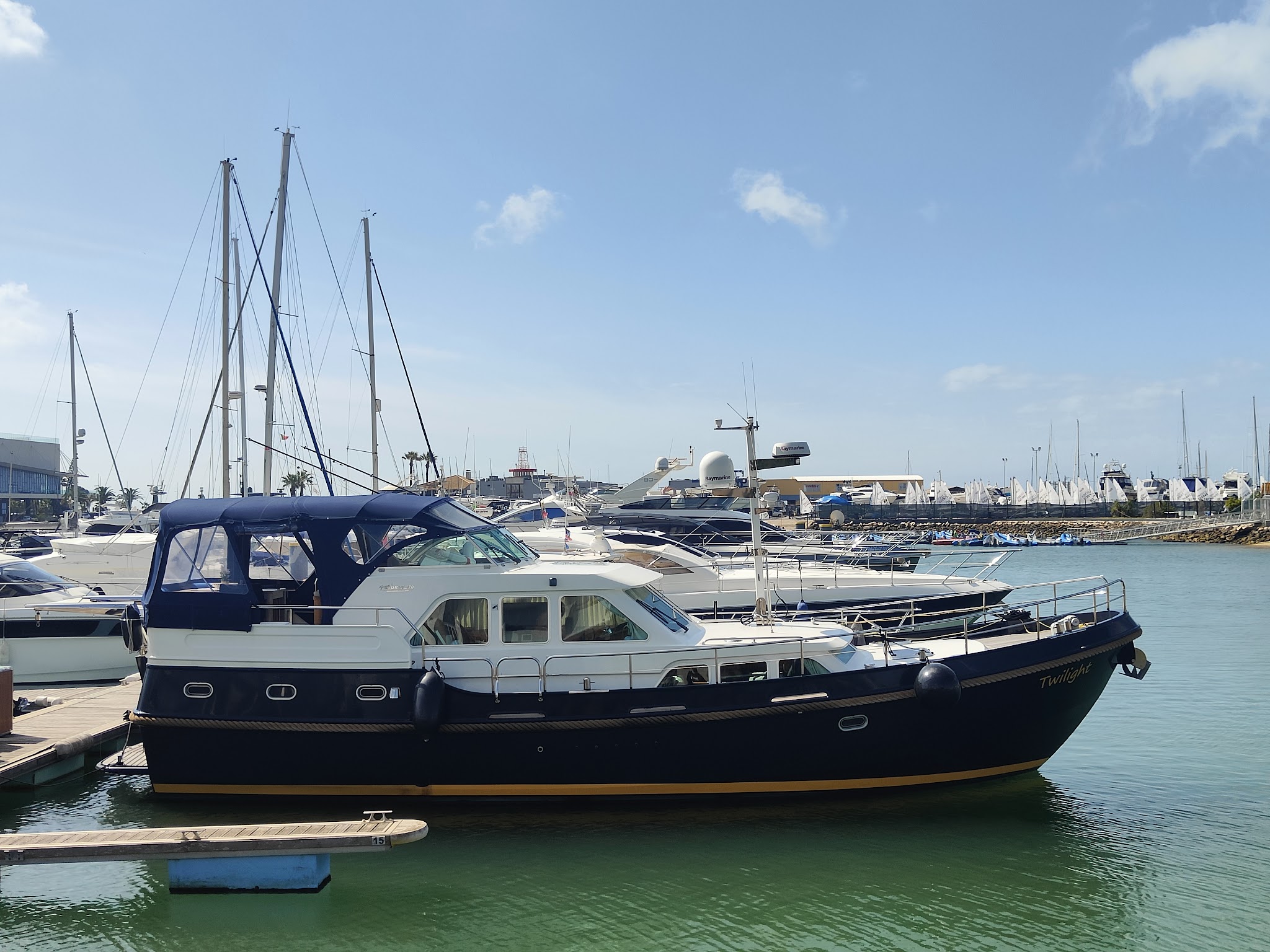 Dunkelblaue Motoryacht liegt am Steg einer sonnigen Marina, umgeben von weiteren Segel- und Motorbooten.