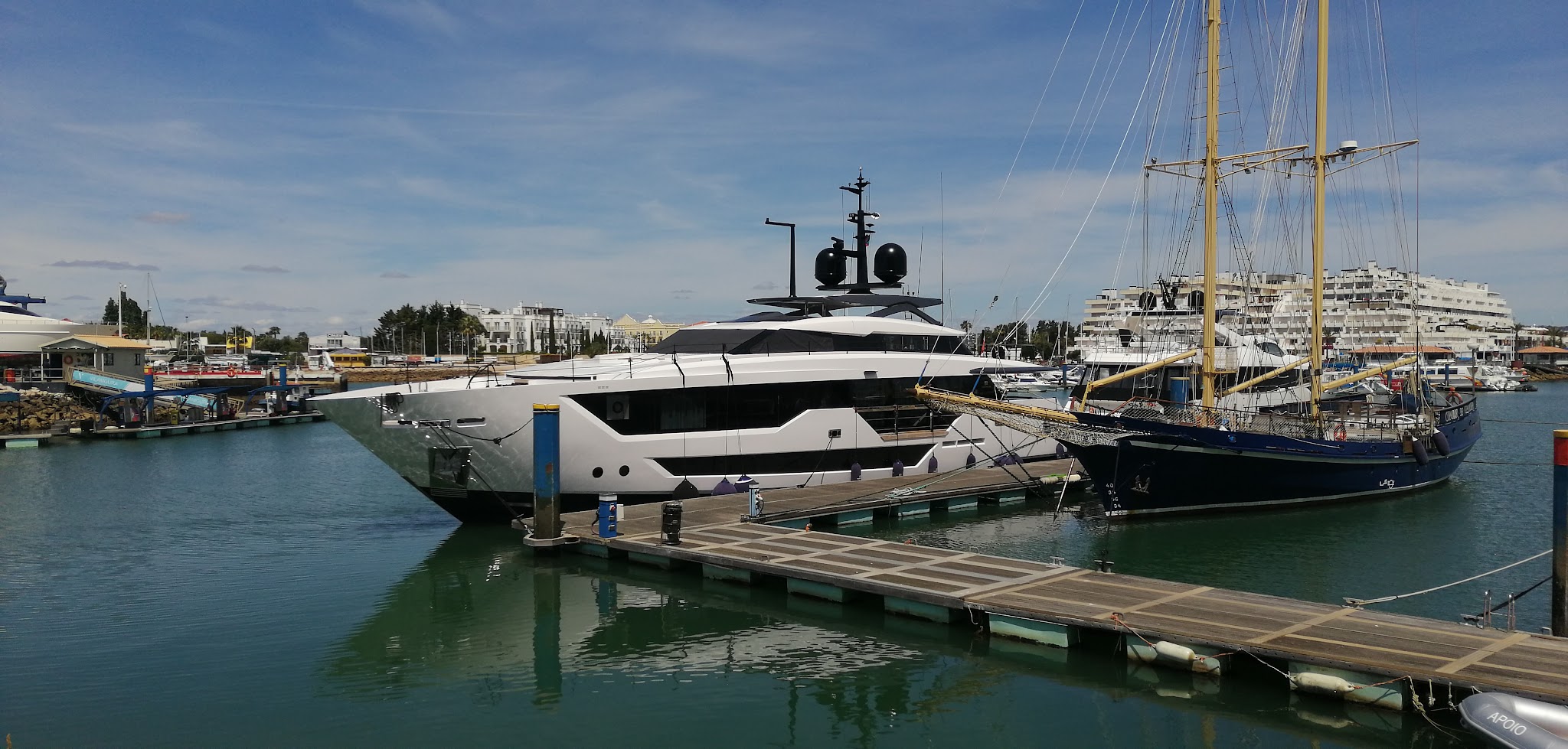 Luxusyacht und Segelschiff liegen an einem sonnigen Hafensteg; moderne Gebäude im Hintergrund, ruhiges Wasser.
