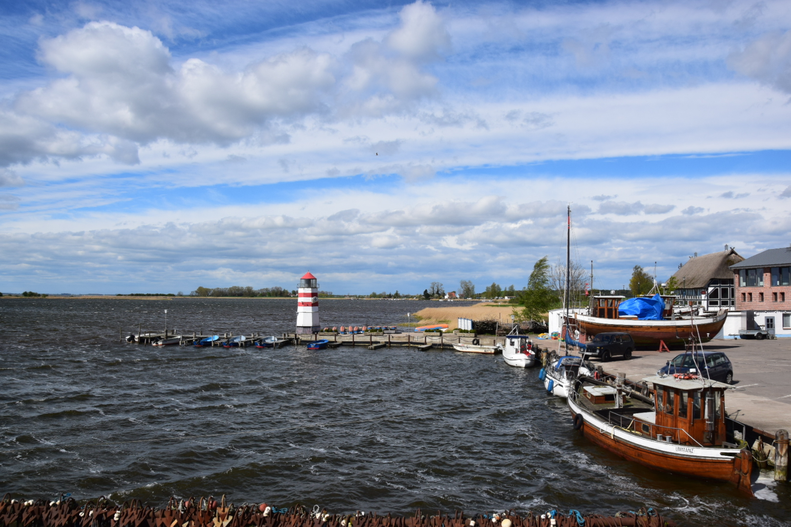 Kleiner Hafen mit rot-weiß gestreiftem Leuchtturm, anliegende Holzboote und welliges Wasser unter teils bewölktem Himmel.