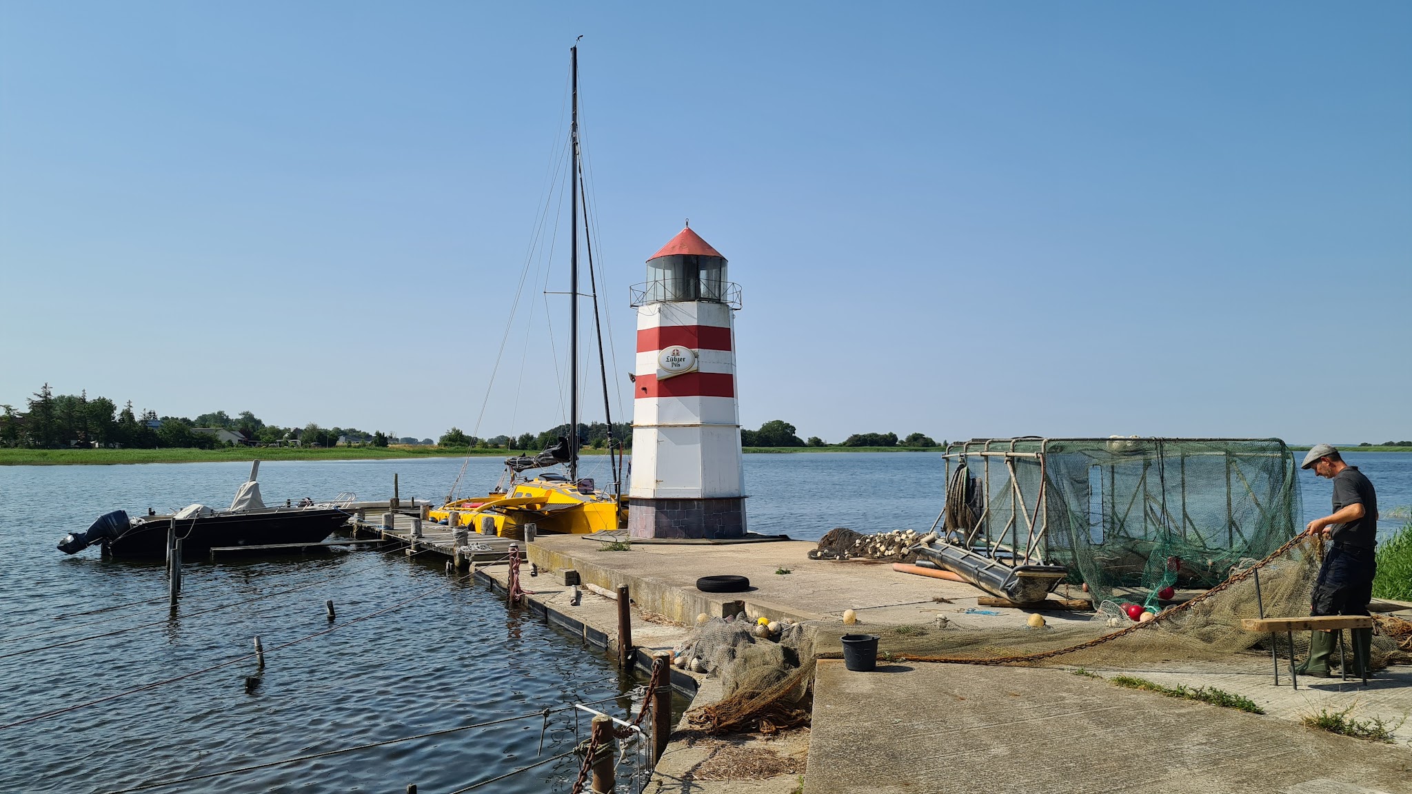 Kleiner rot-weiß gestreifter Leuchtturm auf Steg, daneben Boote; Fischer in Gummistiefeln flickt Netze am sonnigen Seeufer.
