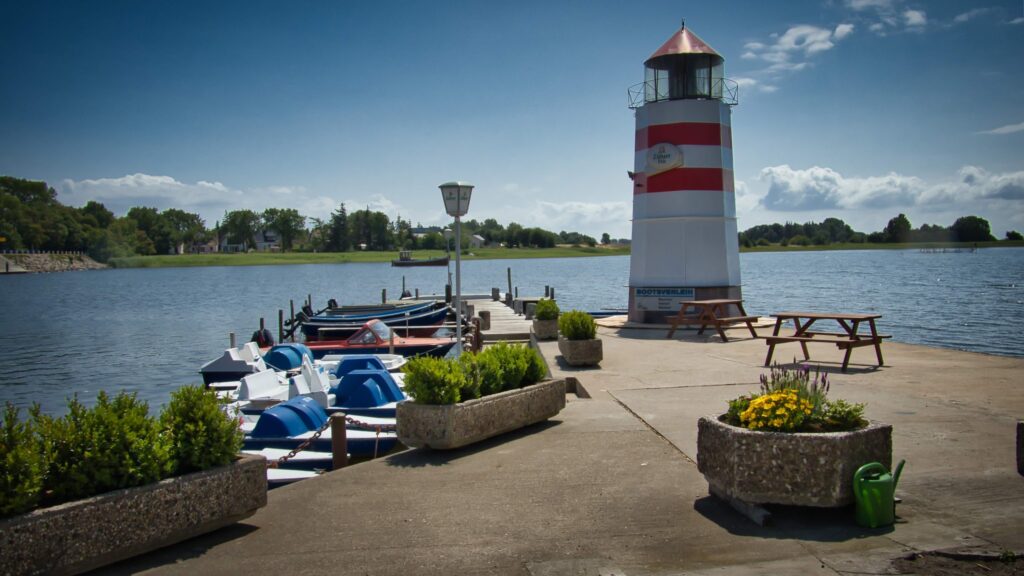 Steg mit Tretbooten, rotem-weißem Leuchtturm und Picknickbänken an ruhigem See unter blauem Himmel