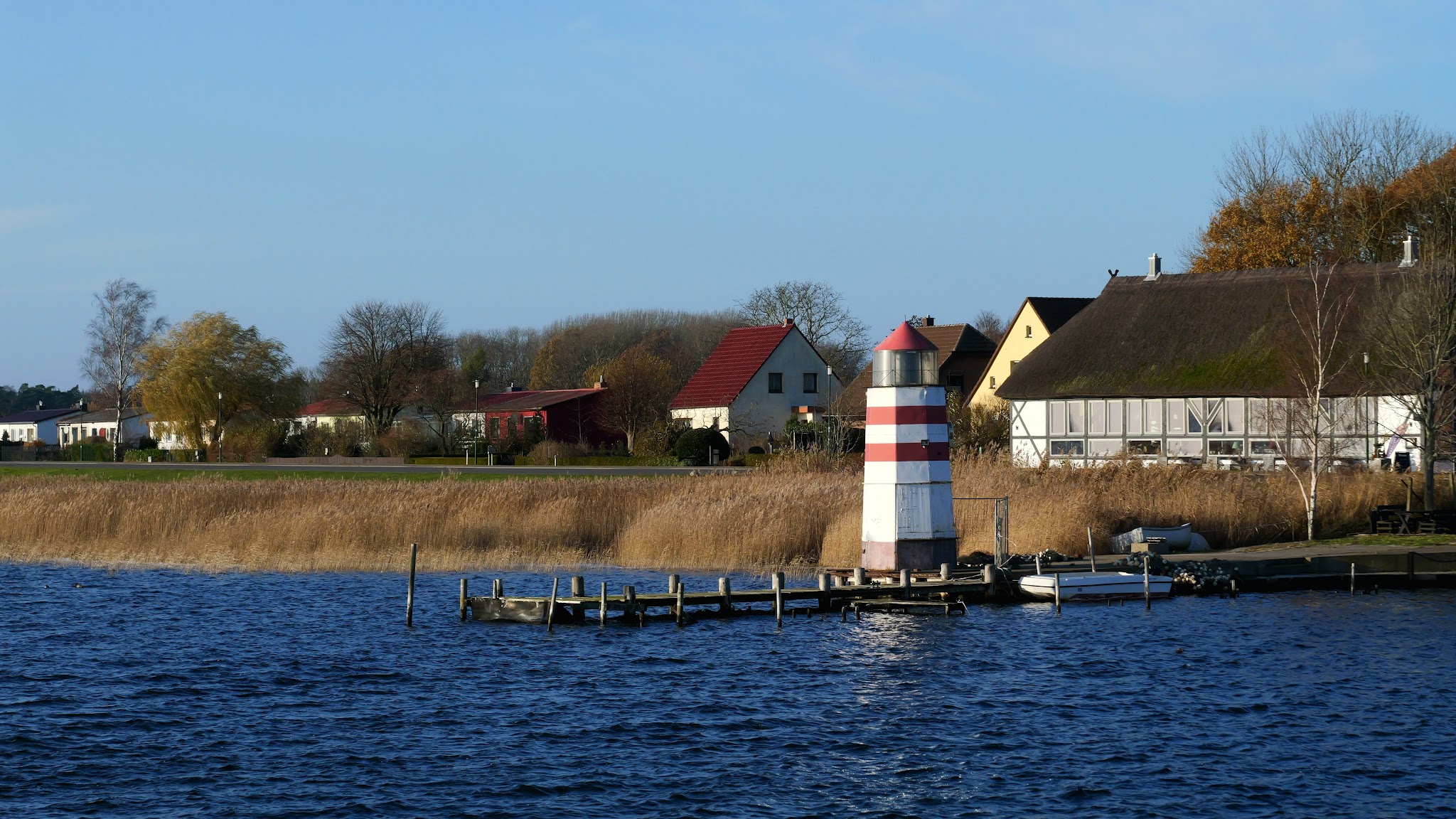 Kleiner rot-weiß gestreifter Leuchtturm am Steg vor Schilf, Dorfhäuser und blauer See unter klarem Himmel