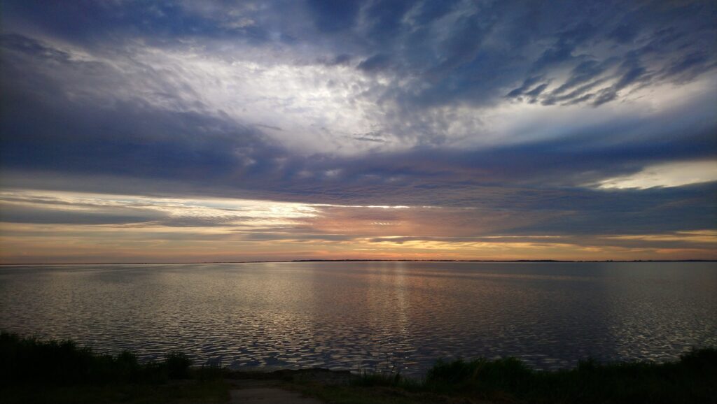 Ruhiger See im Abendlicht, orange-violetter Sonnenuntergang unter dramatischen Wolken am Horizont.