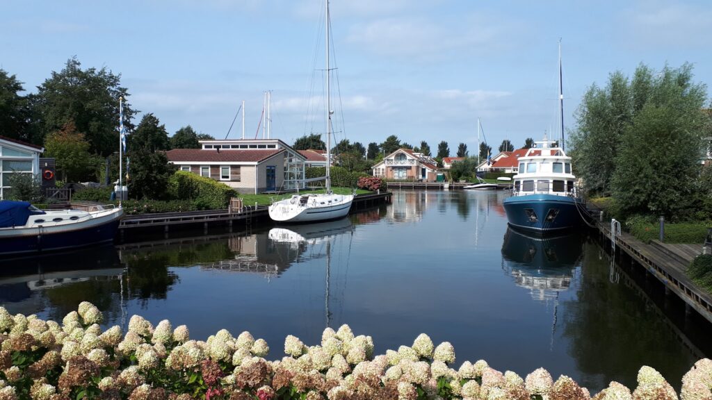 Ruhiger Kanal mit zwei Yachten, Ferienhäusern und weißen Hortensien im Vordergrund unter blauem Himmel.