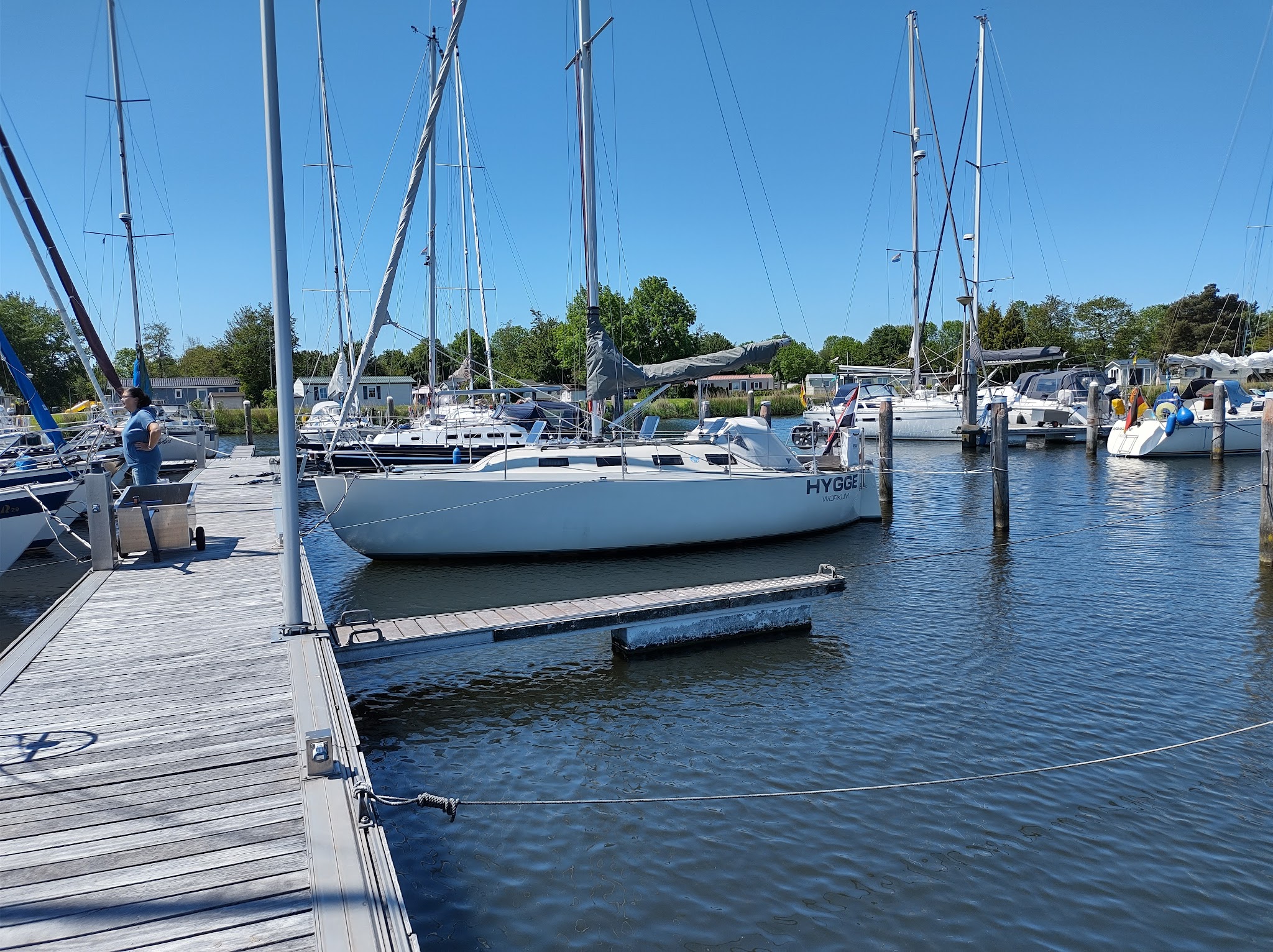 Jachthafen bei Sonnenschein; weißes Segelboot „HYGGE“ längs am Steg, Mann auf dem Holzpier, weitere Boote im Hintergrund.