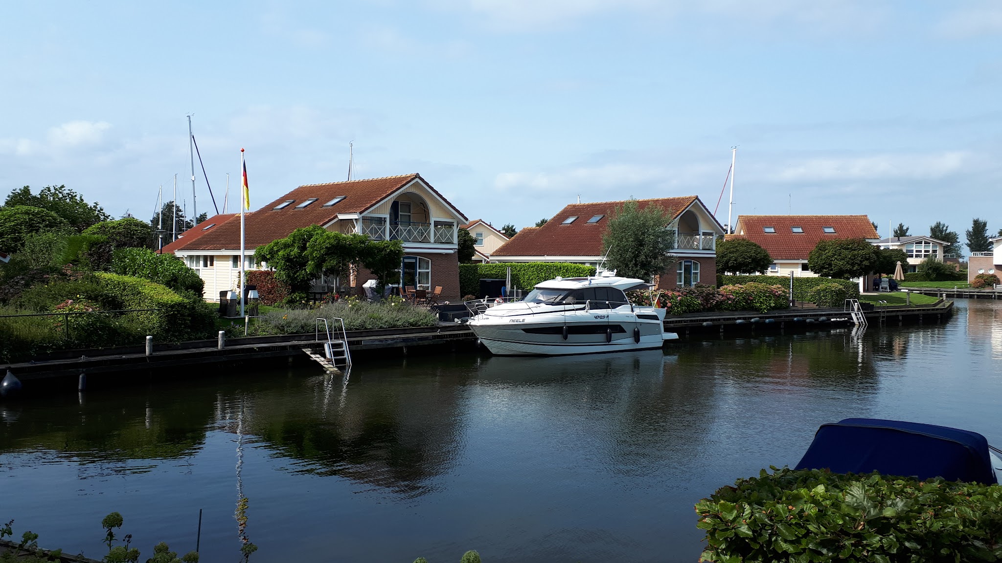Weiße Motoryacht am Steg vor roten Ferienhäusern an ruhigem Kanal; Gärten, Fahnenmast, blauer Himmel.