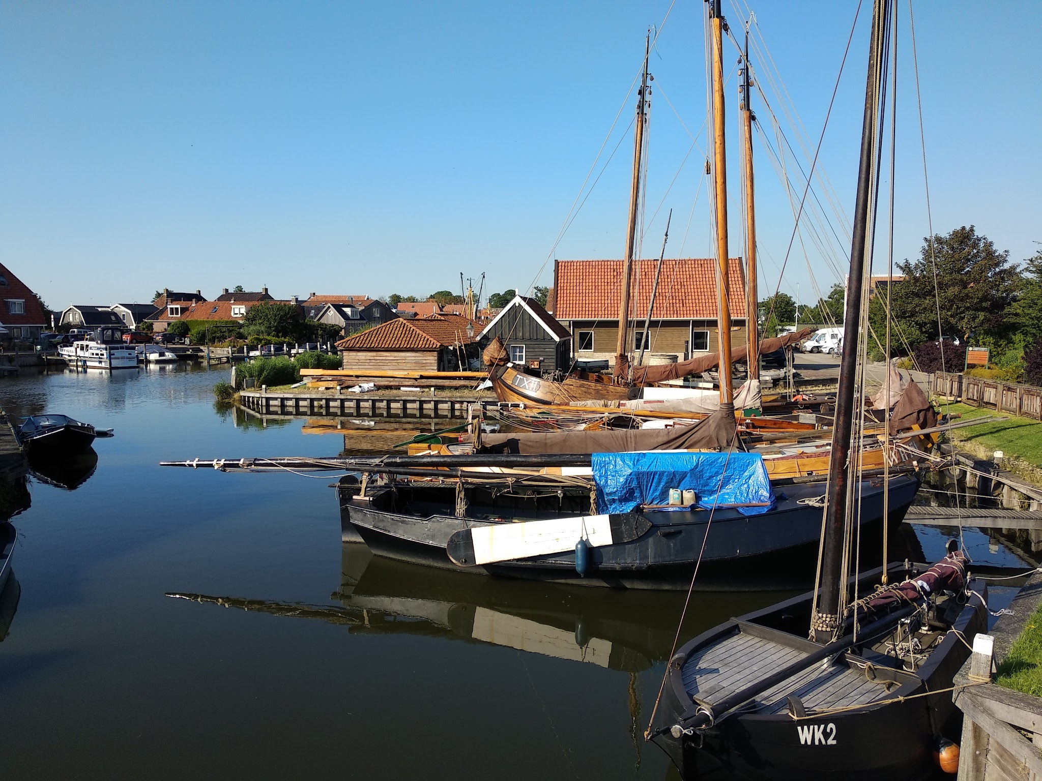 Kleiner Hafen mit mehreren festgemachten historischen Segelbooten, roten Ziegeldach-Häusern im Hintergrund, ruhiges Wasser