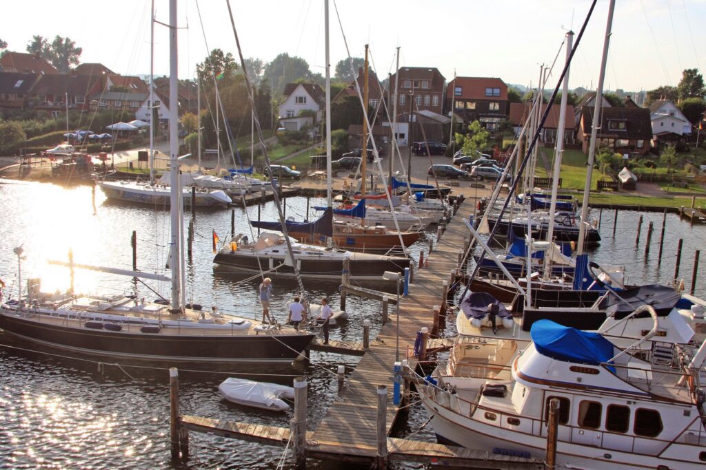 Kleiner Jachthafen: mehrere Segelboote am Steg, Abendsonne spiegelt im Wasser, Menschen hantieren an Bord, Häuser dahinter