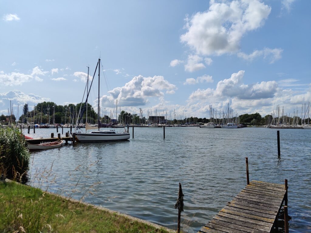 Ruhiger Yachthafen: Segelboote liegen am Steg, kleiner Holzsteg im Vordergrund, blauer Himmel mit Wolken.