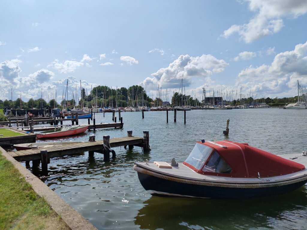 Kleines Motorboot mit rotem Verdeck am Steg, im Hintergrund Yachthafen voller Segelboote unter blauem Himmel.