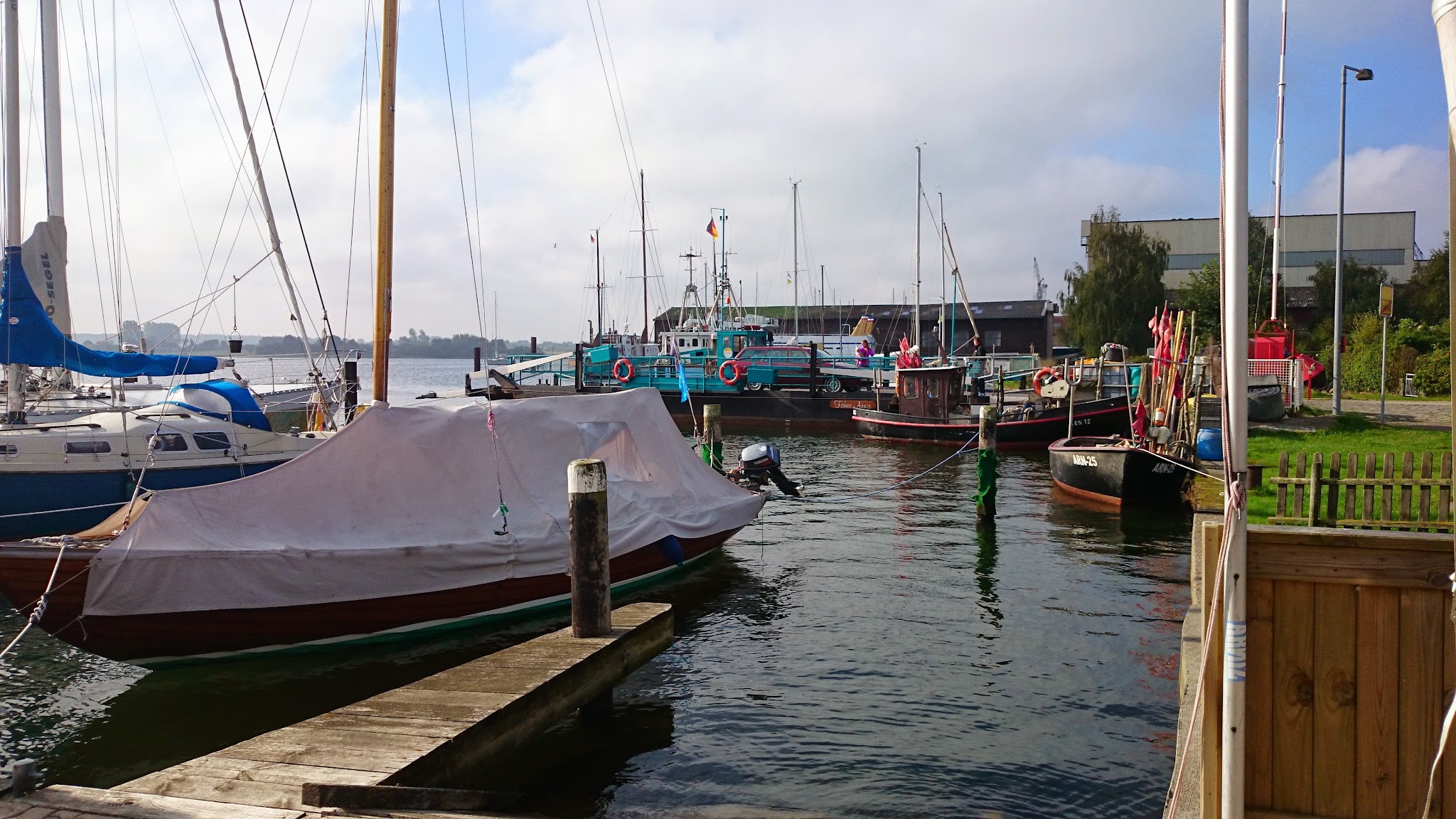 Ruhiger Hafen: bedecktes Segelboot am Steg, mehrere bunte Fischer- und Segelschiffe vor bewölktem Himmel