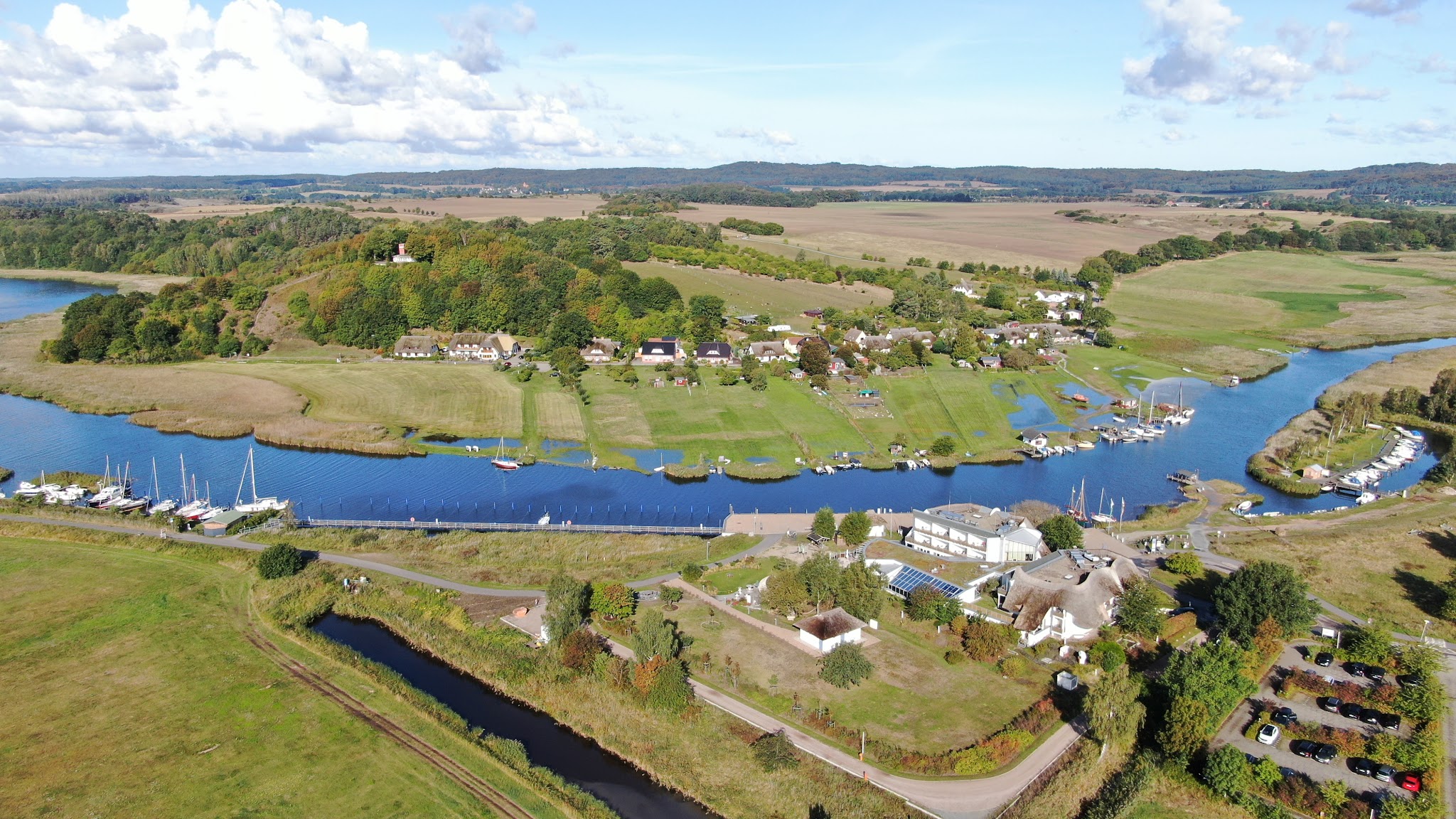Luftaufnahme: Fluss mit Yachthafen und Segelbooten, Dorfhäuser, Felder und bewaldete Hügel im Sonnenschein.