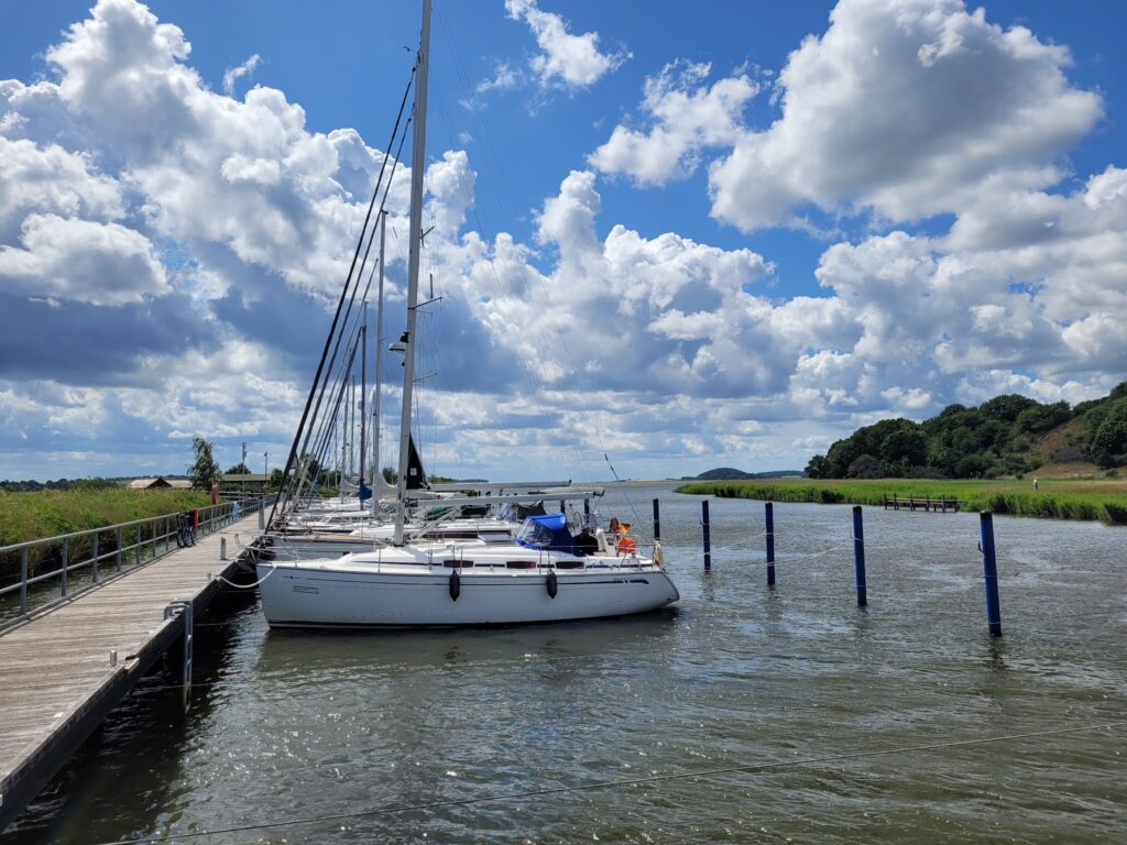 Segelboote liegen an einem Holzsteg, davor ruhiges Wasser; im Hintergrund grüne Ufer und wolkiger Sommerhimmel