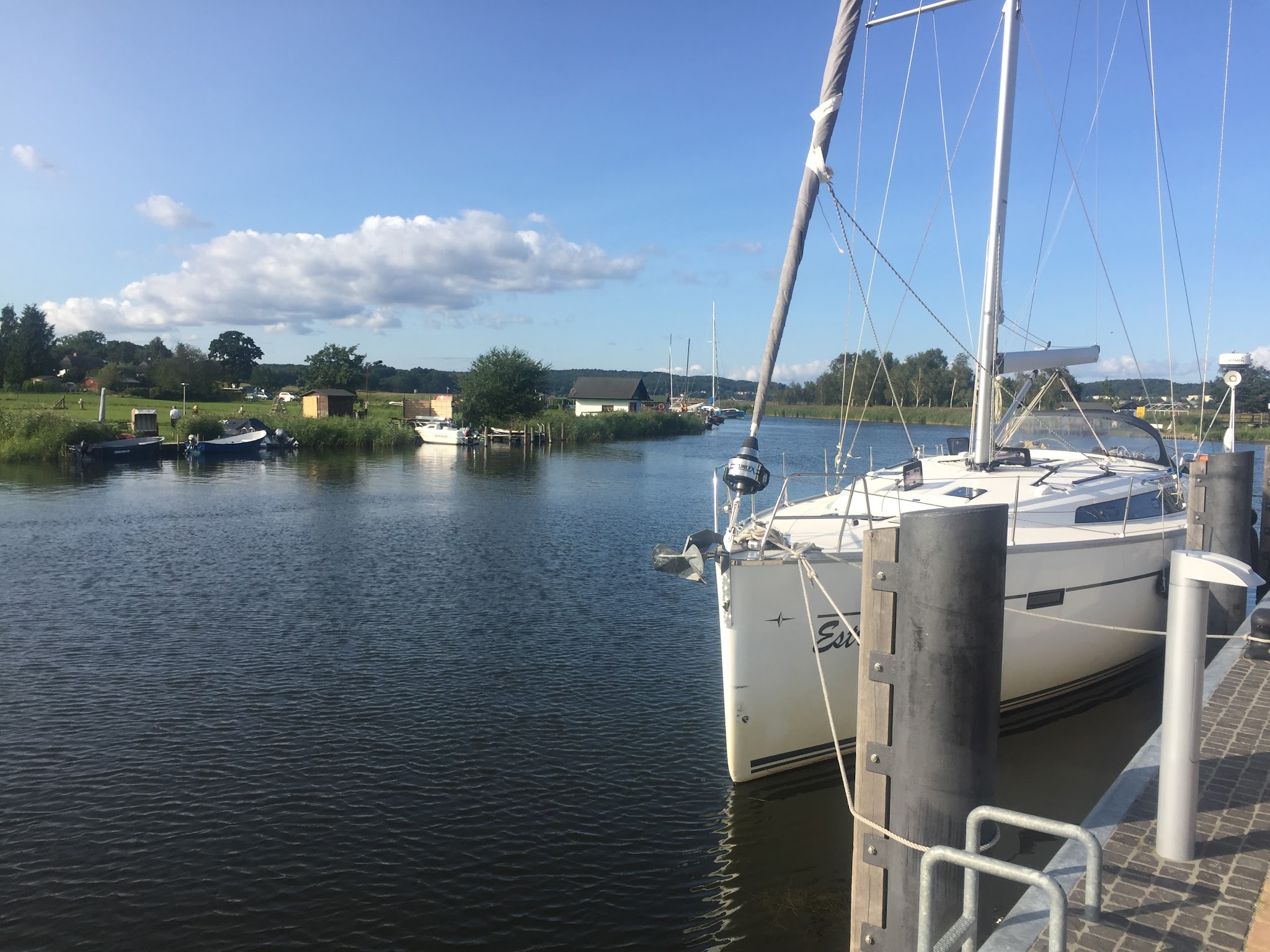 Segelboot am Steg auf ruhigem Fluss, grüne Ufer mit kleinen Booten, blauer Himmel mit vereinzelten Wolken