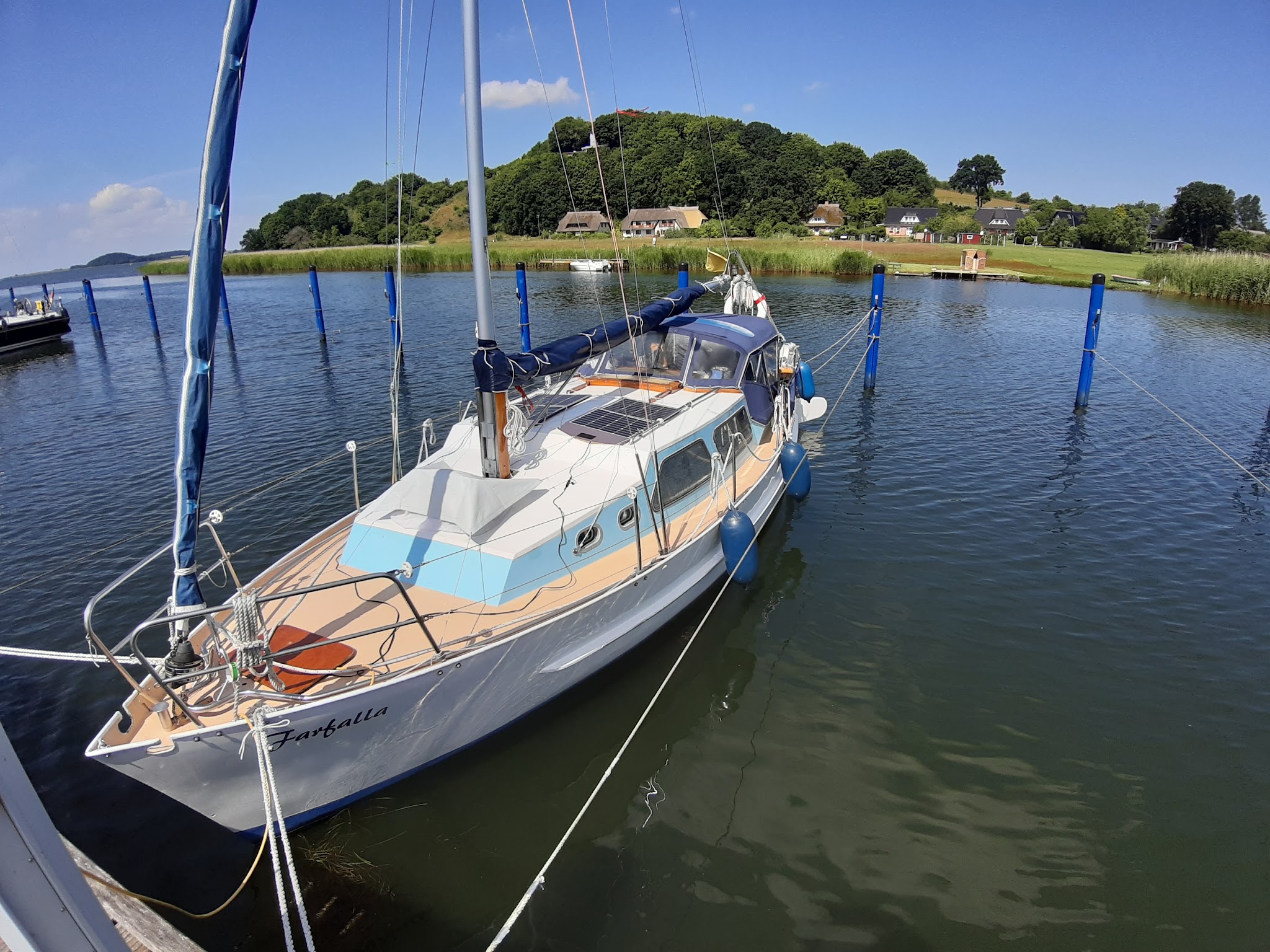 Kleines Segelboot im ruhigen Hafen, vertäut zwischen blauen Pfosten; grüner Hügel mit Häusern unter blauem Himmel.