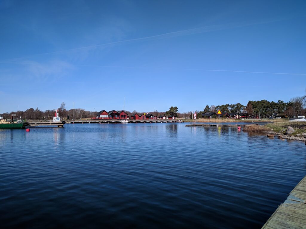 Ruhiger Hafen, rote Bootshäuser an Stegen, spiegelndes Wasser unter klarem blauen Himmel.