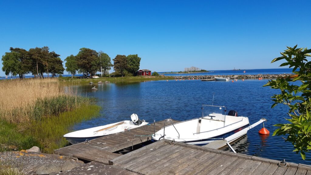 Zwei kleine Motorboote an Holzsteg, ruhige Bucht mit Schilf, rotem Hüttchen und Bäumen unter klarem Himmel