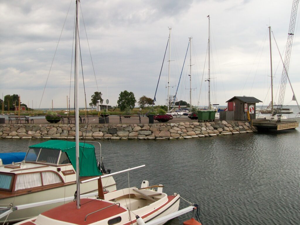 Kleiner Hafen mit mehreren Segelbooten an einem Steinpier unter bewölktem Himmel, ruhiges Wasser im Vordergrund
