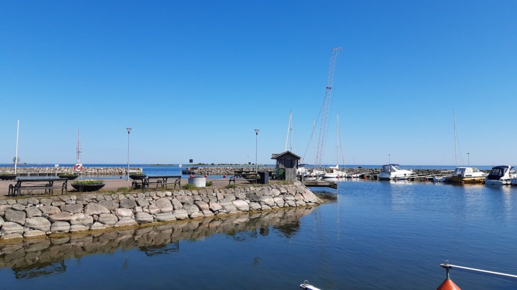 Kleiner Yachthafen: Segel- und Motorboote an Stegen, Holzhütte, Steinmole, ruhiges Wasser unter wolkenlosem Himmel