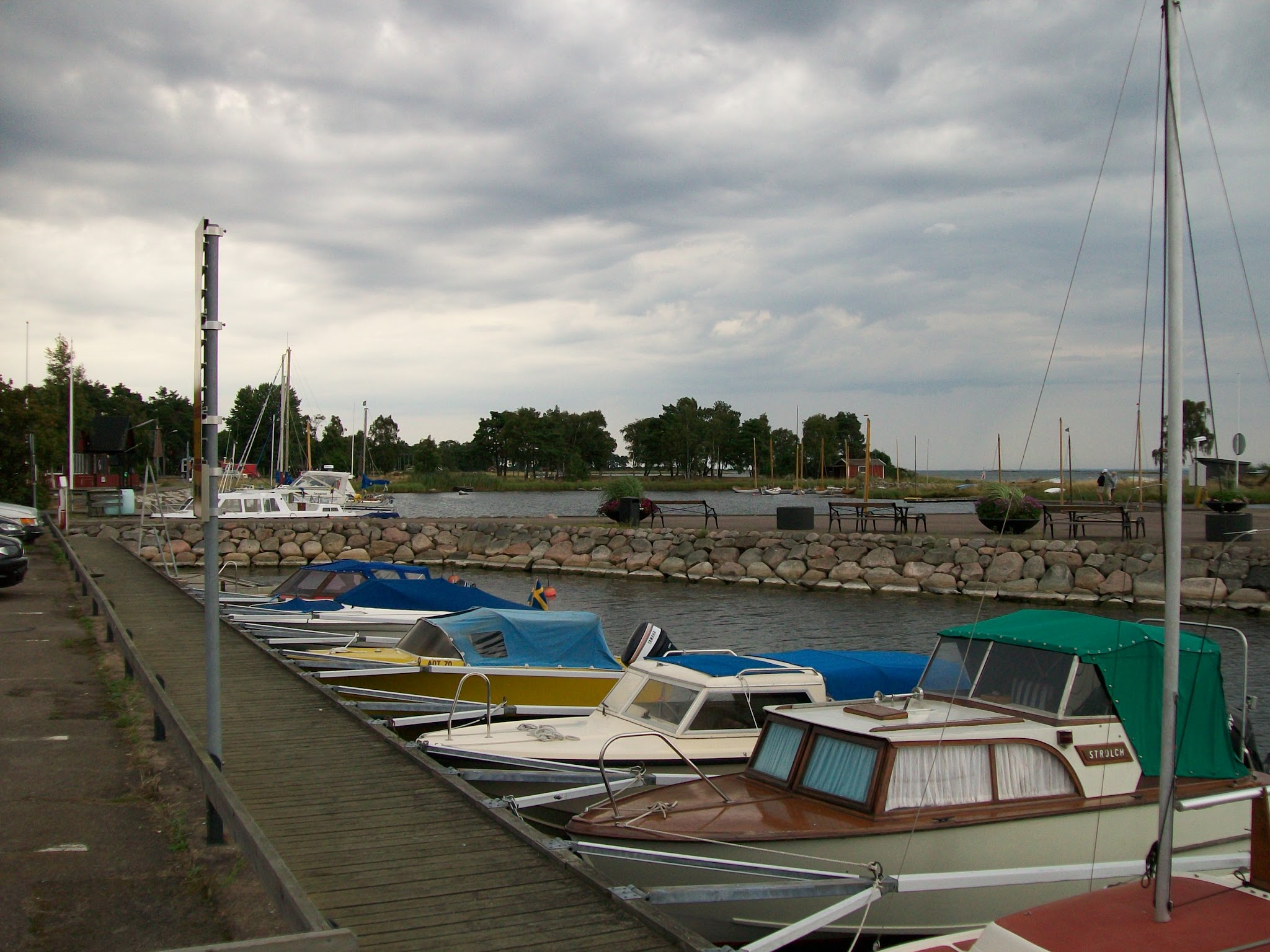 Kleine Motorboote liegen am Steg eines Jachthafens; dahinter Steinmauer, Bäume und wolkenverhangener Himmel.