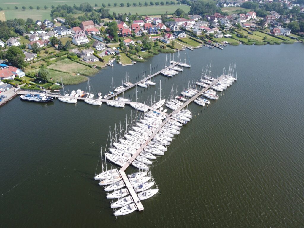 Luftaufnahme eines Yachthafens voller Segelboote vor einer kleinen Ortschaft am Seeufer
