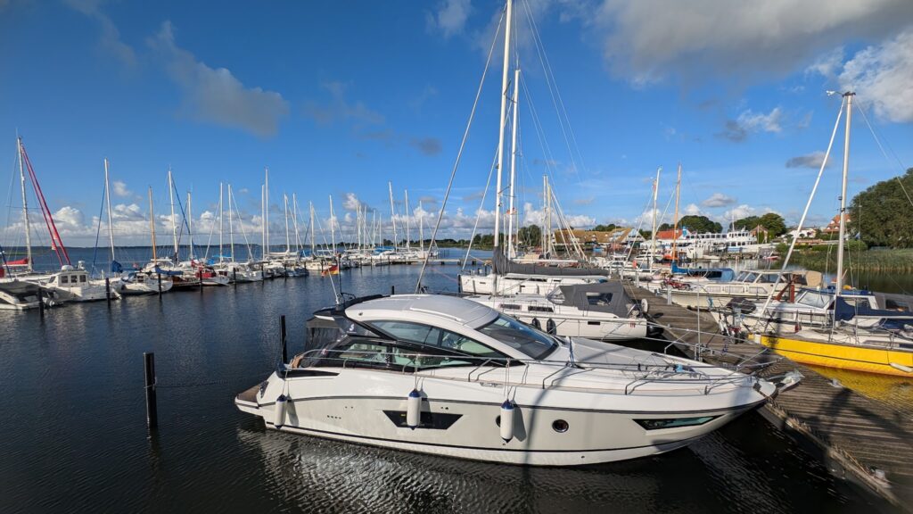 Weißes Motorboot im Yachthafen, umgeben von zahlreichen Segelbooten bei sonnigem Wetter unter blauem Himmel.