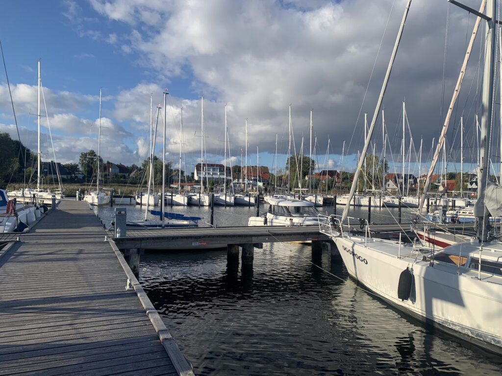Holzsteg in kleinem Jachthafen mit zahlreichen vertäuten Segelbooten unter teils bewölktem Himmel