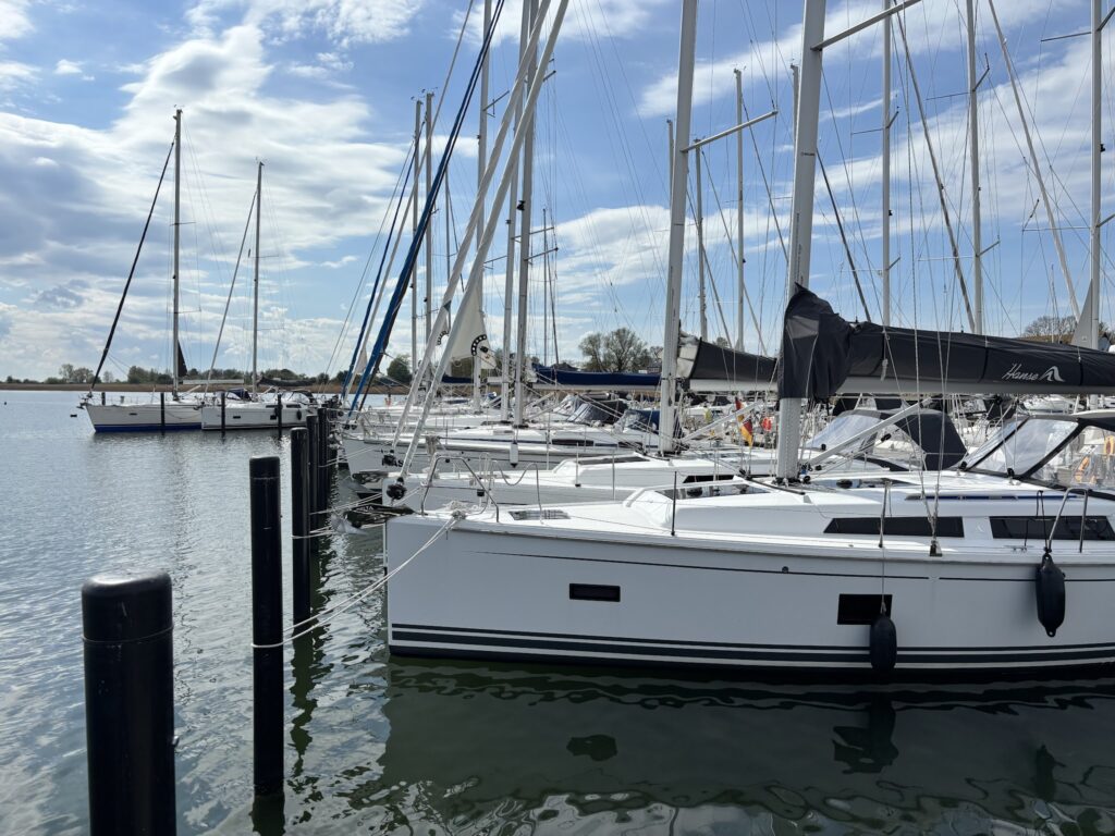 Reihe weißer Segelyachten mit hohen Masten in ruhigem Hafen; spiegelndes Wasser, teils bewölkter blauer Himmel