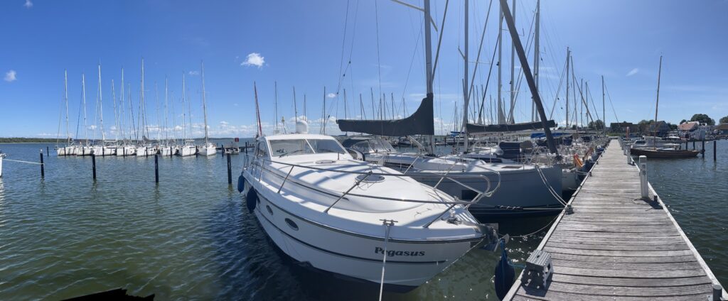 Yachten und Segelboote liegen bei Sonnenschein am Holzsteg eines ruhigen Hafens, blauer Himmel spiegelt im Wasser.