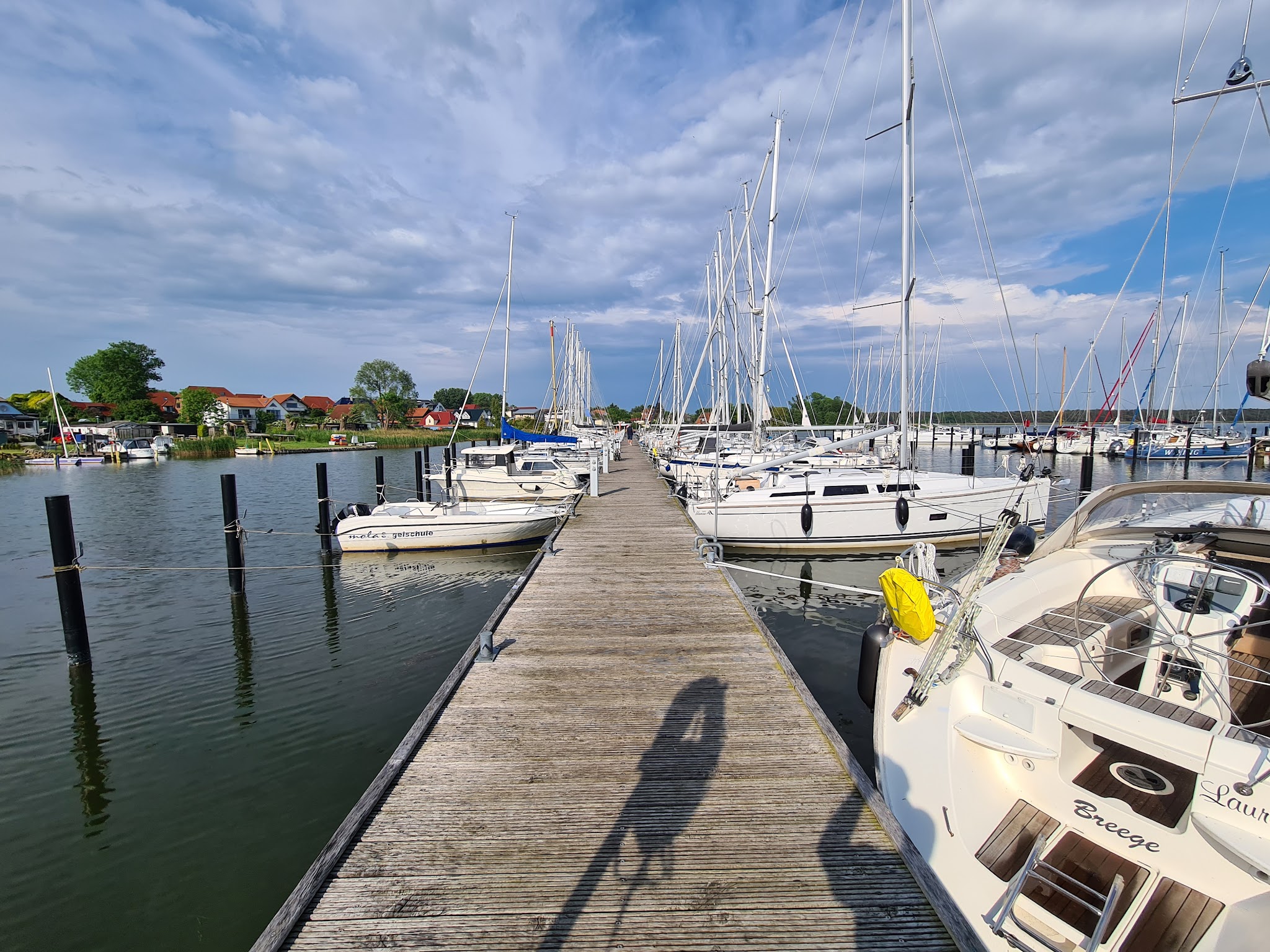 Holzsteg mit vielen festgemachten Segelbooten im Hafen, ruhiges Wasser, im Hintergrund Häuser und wolkiger Himmel.