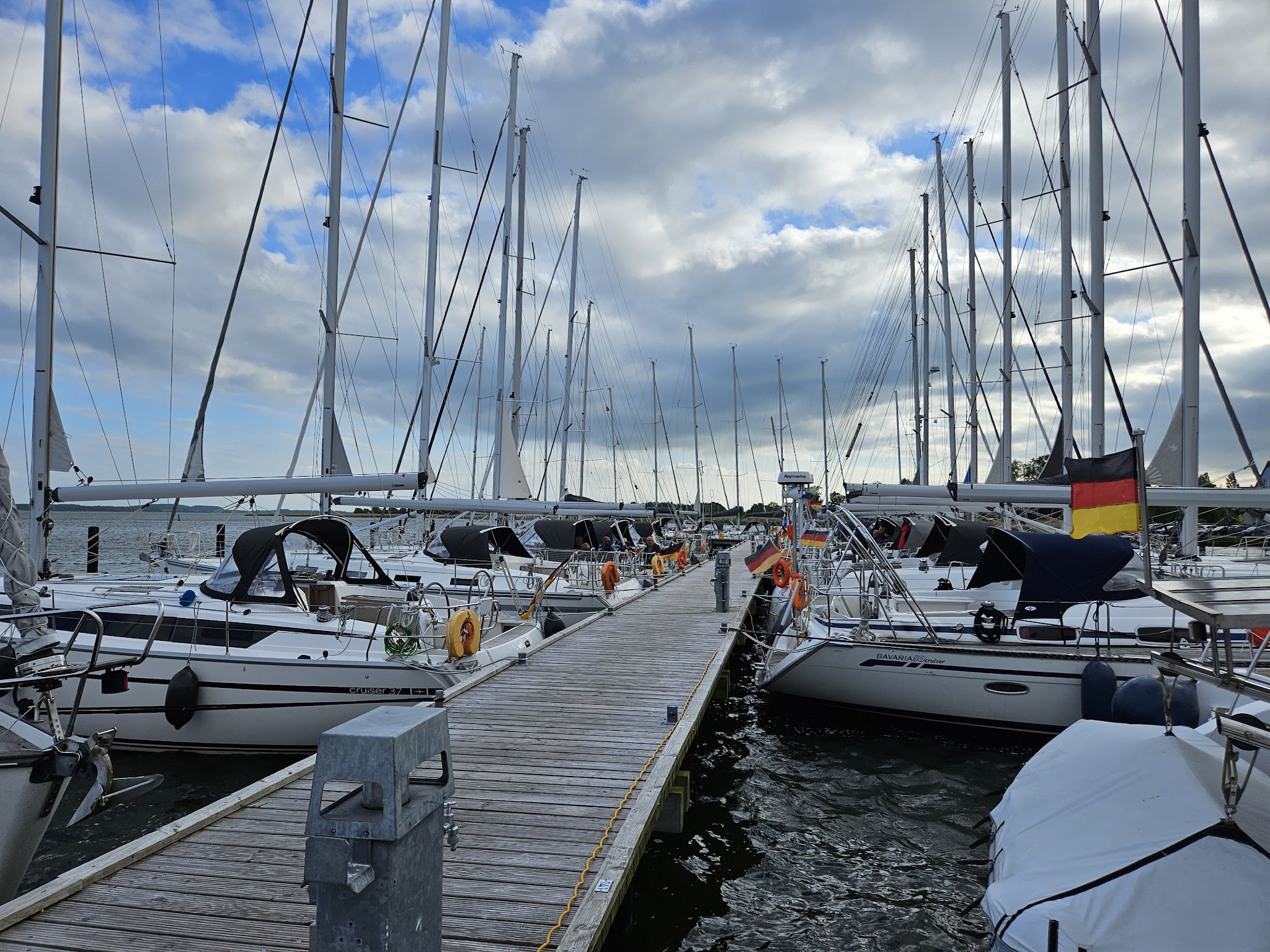 Segelyachten mit hohen Masten liegen beidseitig an einem Holzsteg im Hafen, deutsche Flagge im Wind, wolkiger Himmel.